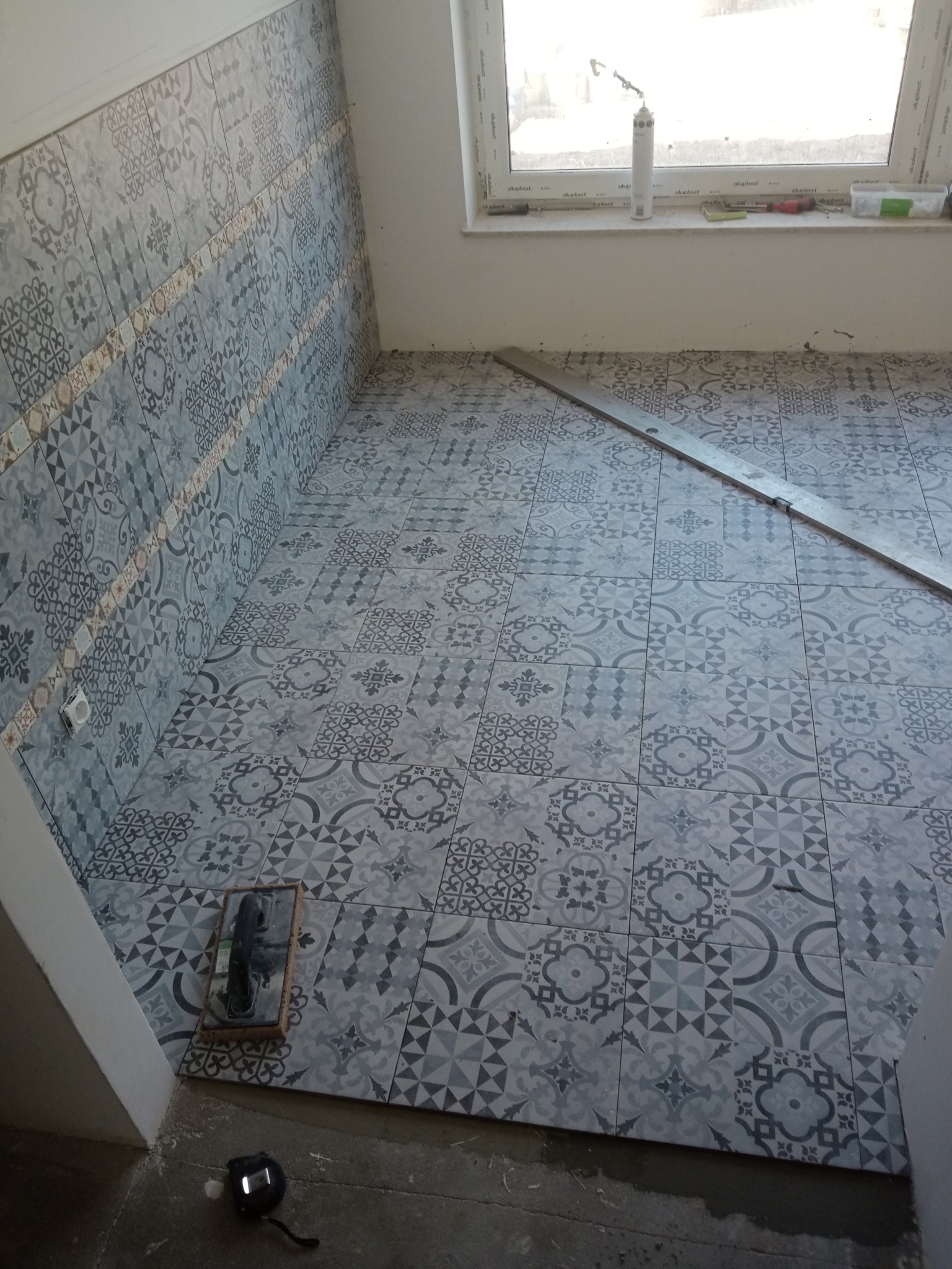 Układanie szaro-białych płytek podłogowych i ściennych z wzorem patchworkowym w trakcie remontu łazienki, widoczna poziomica, paca i miara.