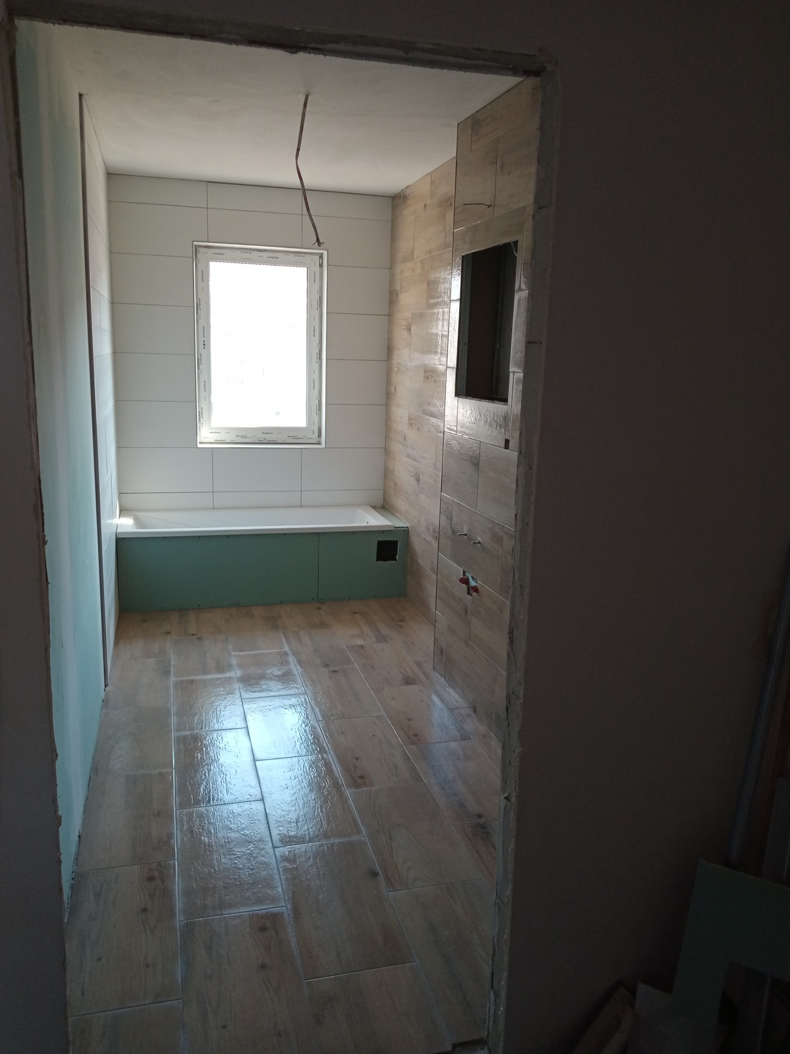 Wnętrze łazienki w trakcie remontu w Kołczygłowach: widoczne płytki imitujące drewno na podłodze, białe płytki na ścianach, wanna w obudowie z zielonych płyt oraz fragment ściany z drewnopodobnymi...