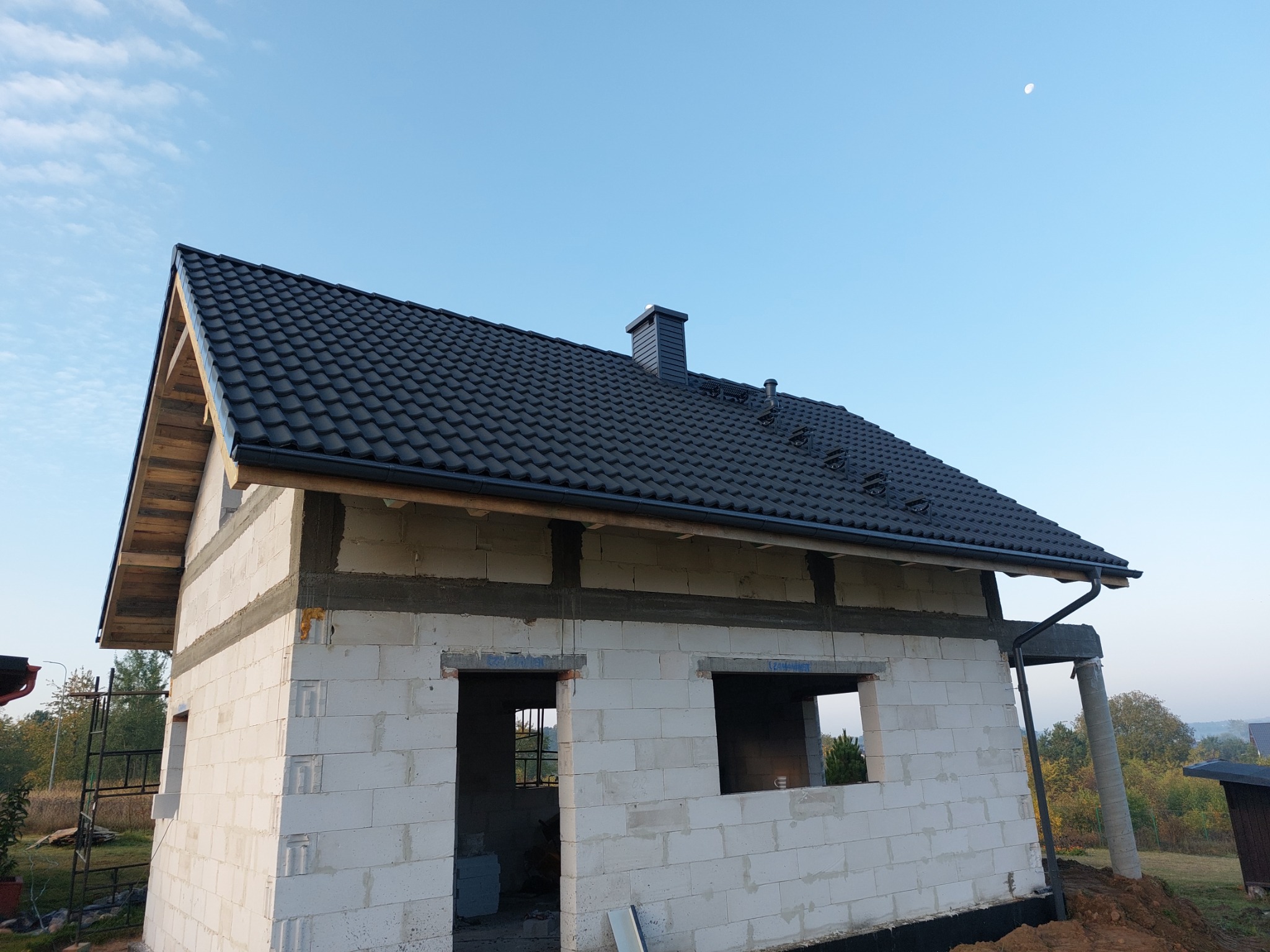 Surowy stan domu z białych bloczków, z ciemną dachówką i widocznym kominem, pod błękitnym niebem z delikatnymi chmurami i księżycem.