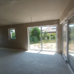 Surowe wnętrze domu jednorodzinnego w trakcie prac wykończeniowych, widoczne ściany i sufit po nałożeniu wstępnej warstwy tynku, z wystającymi przewodami elektrycznymi z sufitu i dużymi oknami...