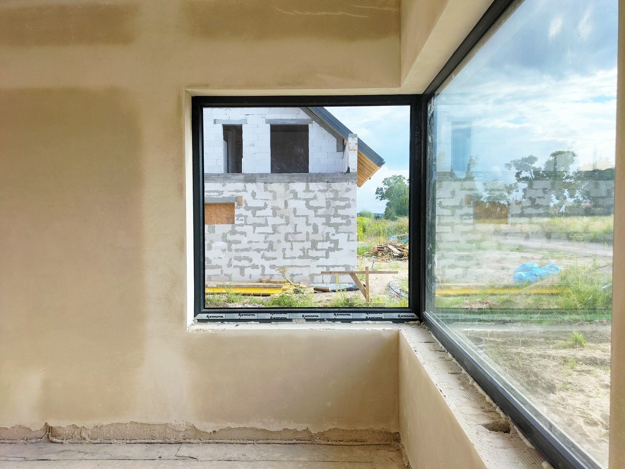 Ściana wewnętrzna w trakcie prac wykończeniowych, widoczne nierówności na powierzchni, przez okno widać budynek w stanie surowym otwartym i zieleń.