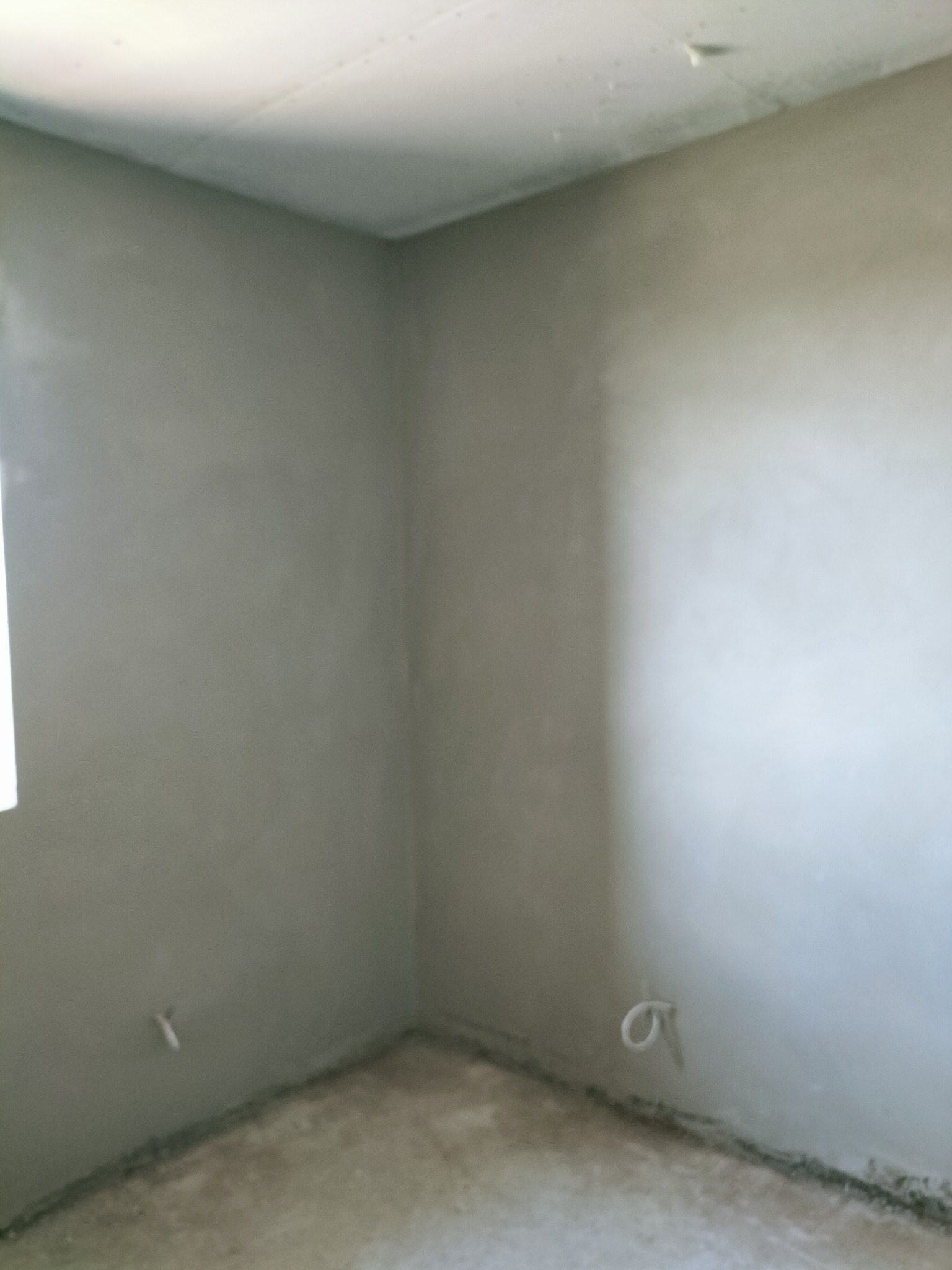 Wnętrze pokoju po nałożeniu świeżej, szarej warstwy wykończeniowej na ścianach i suficie, widoczne wystające kable elektryczne.