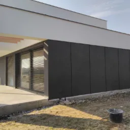 Nowoczesny dom z elewacją w odcieniach szarości i bieli, widoczne rolety zewnętrzne, teren wok&oacute;ł domu w trakcie zagospodarowywania, z elementami budowlanymi i wiaderkiem.