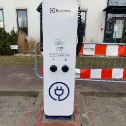 Biała stacja ładowania samochodów elektrycznych marki Electrolux z dwoma gniazdami, umieszczona na zewnątrz budynku biurowego, zabezpieczona barierkami tymczasowymi z napisem Salvada.