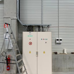 Time to Start (Danylo Chernytsia) - Biała szafa sterownicza z zielonymi lampkami kontrolnymi i symbolami ostrzegawczymi, zamontowana w hali przemysłowej z widoczną drabinką, gaśnicą i instalacją elektryczną pod sufitem.