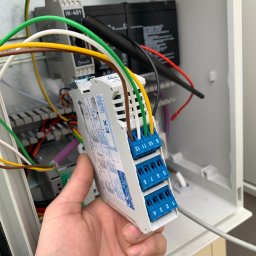 Time to Start (Danylo Chernytsia) - Ręka trzyma moduł sterujący z niebieskimi złączami i podłączonymi kolorowymi przewodami wewnątrz skrzynki elektrycznej, widoczne akumulatory i inne elementy instalacji.