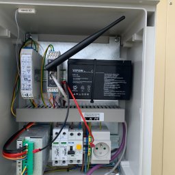 Time to Start (Danylo Chernytsia) - Wnętrze skrzynki elektrycznej z akumulatorem VIPOW, modułami SIMTEC, zasilaczem, gniazdkiem i anteną WiFi, okablowanie uporządkowane, widoczne elementy instalacji alarmowej lub zasilania awaryjnego.