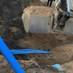 Time to Start (Danylo Chernytsia) - Wykop pod fundamenty budynku z niebieską rurą karbowaną ułożoną wzdłuż ściany, łyżka koparki w trakcie nabierania ziemi.