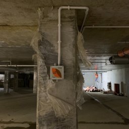 Time to Start (Danylo Chernytsia) - Słup w surowym stanie, zabezpieczony folią, z zamontowaną skrzynką elektryczną z logo, białe rury instalacyjne, w tle pracownik na drabinie w pomarańczowej kamizelce, instalacja w trakcie...