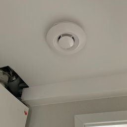 Time to Start (Danylo Chernytsia) - Biały okrągły czujnik dymu zamontowany na białym suficie, widoczne fragmenty instalacji wentylacyjnej i ściany z szarą mozaiką.