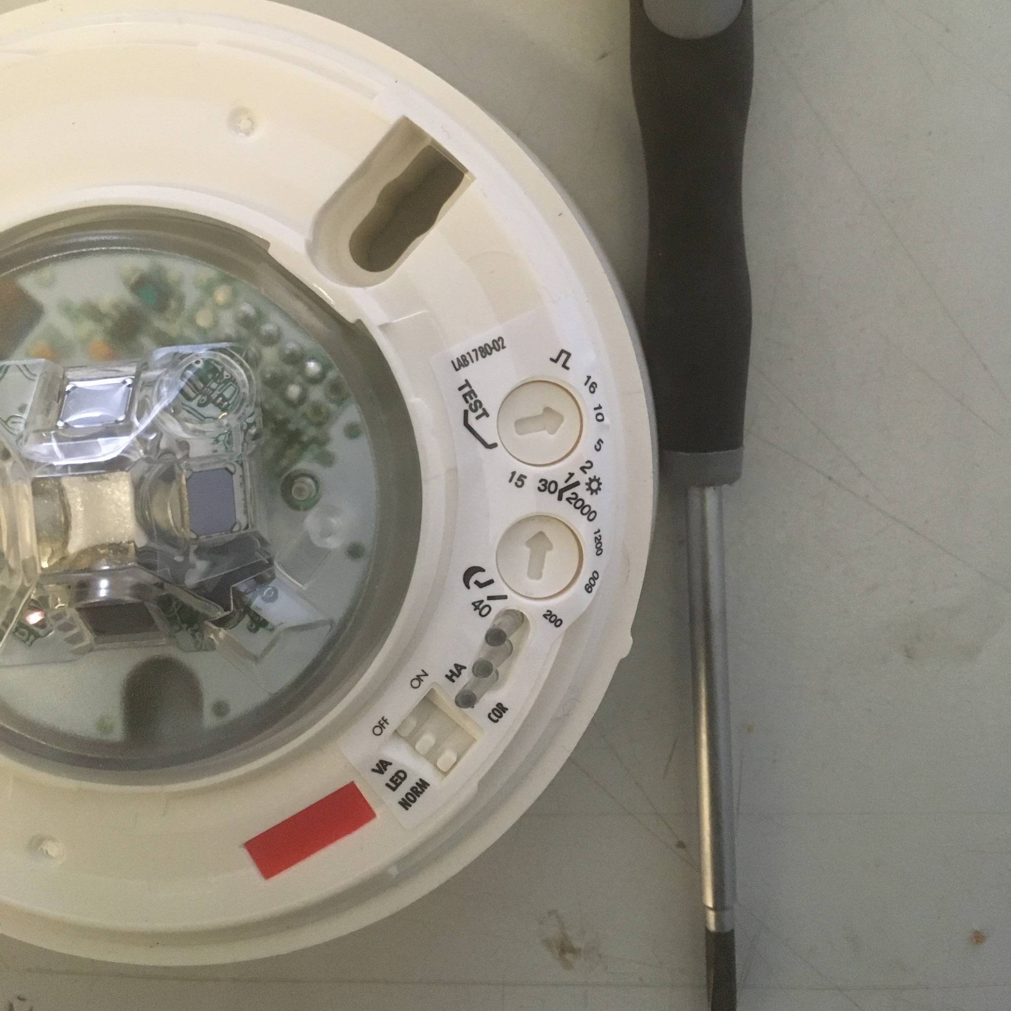 Otwarty czujnik dymu z widocznymi elementami elektronicznymi i ustawieniami, obok leży śrubokręt.