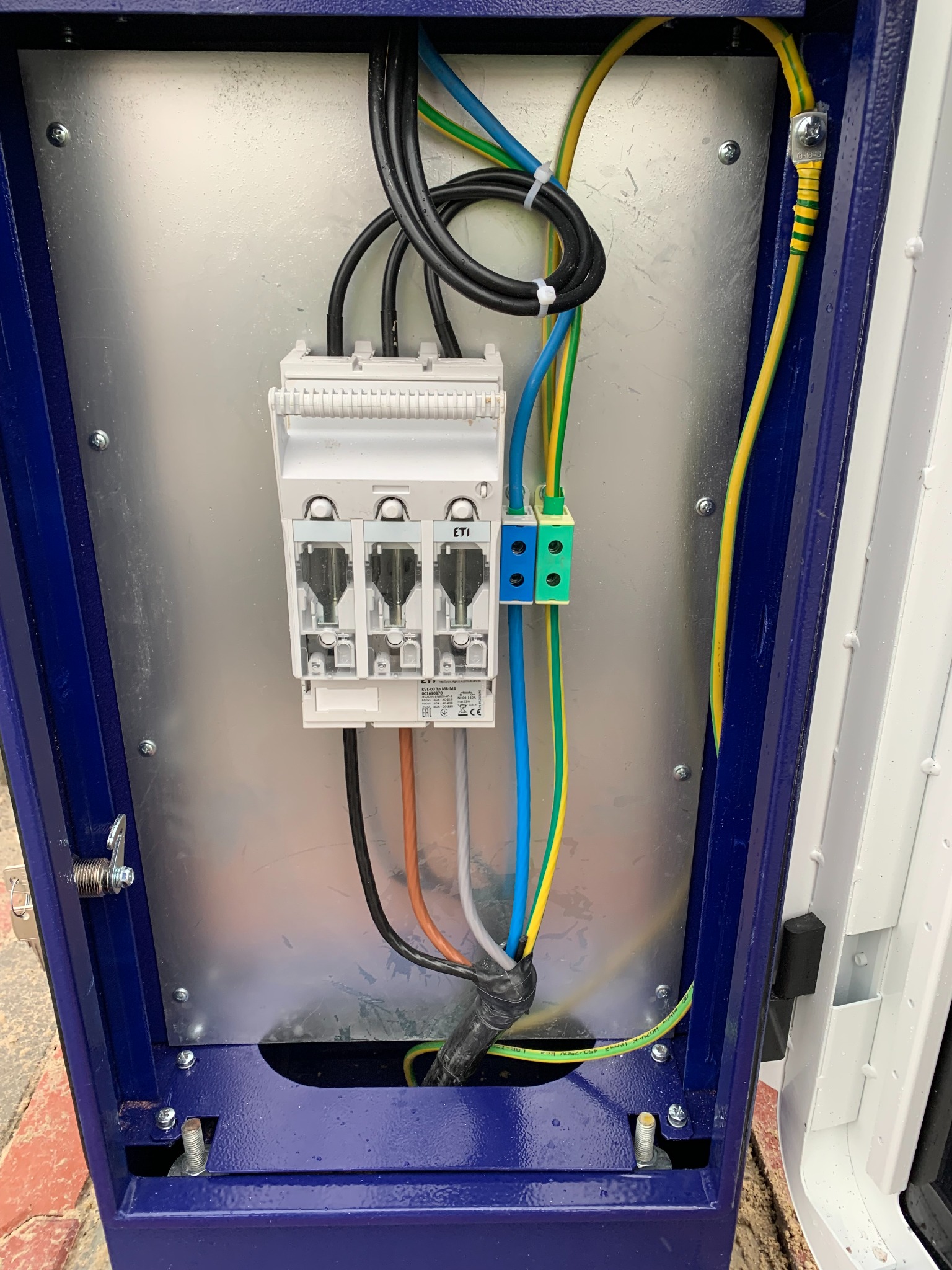Wnętrze skrzynki elektrycznej z zamontowanym bezpiecznikiem ETI, przewodami w kolorach: czarnym, zielono-żółtym, niebieskim i brązowym, oraz złączkami w kolorach niebieskim i zielonym.