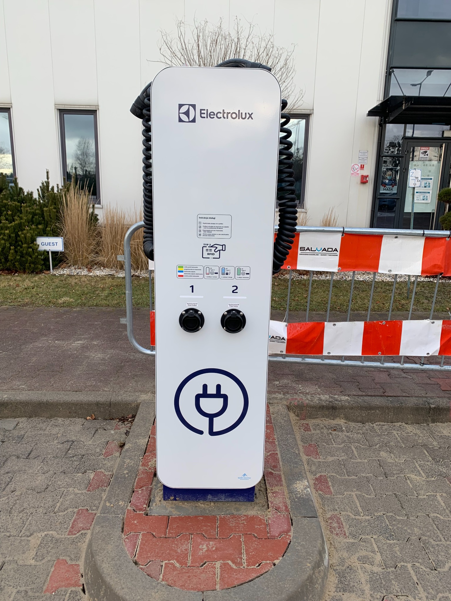 Biała stacja ładowania samochodów elektrycznych marki Electrolux z dwoma gniazdami, umieszczona na zewnątrz budynku biurowego, zabezpieczona barierkami tymczasowymi z napisem Salvada.