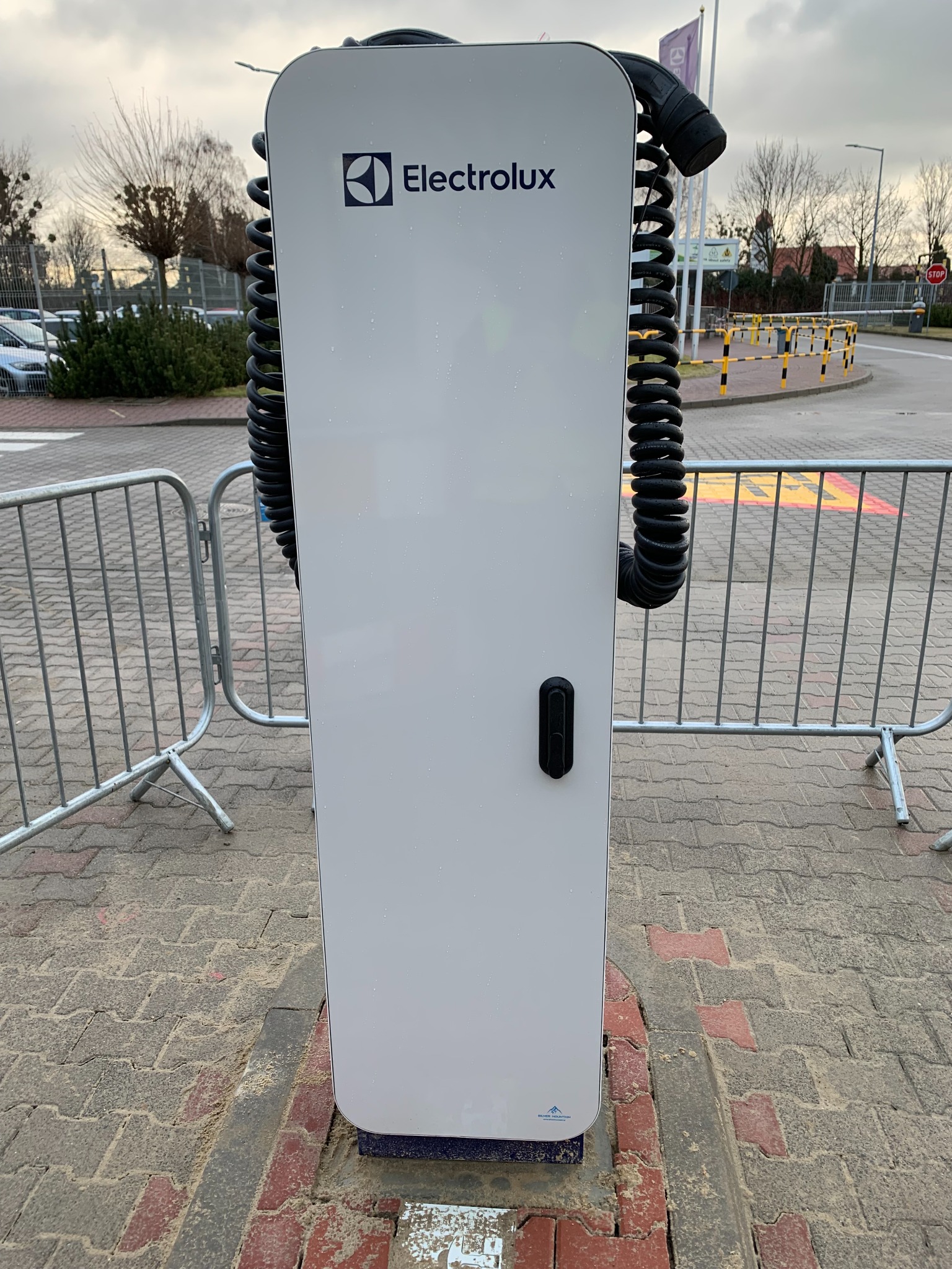 Biała stacja ładowania pojazdów elektrycznych marki Electrolux z czarnym, spiralnym kablem, umieszczona na kostce brukowej w otoczeniu metalowych barierek i elementów infrastruktury drogowej.