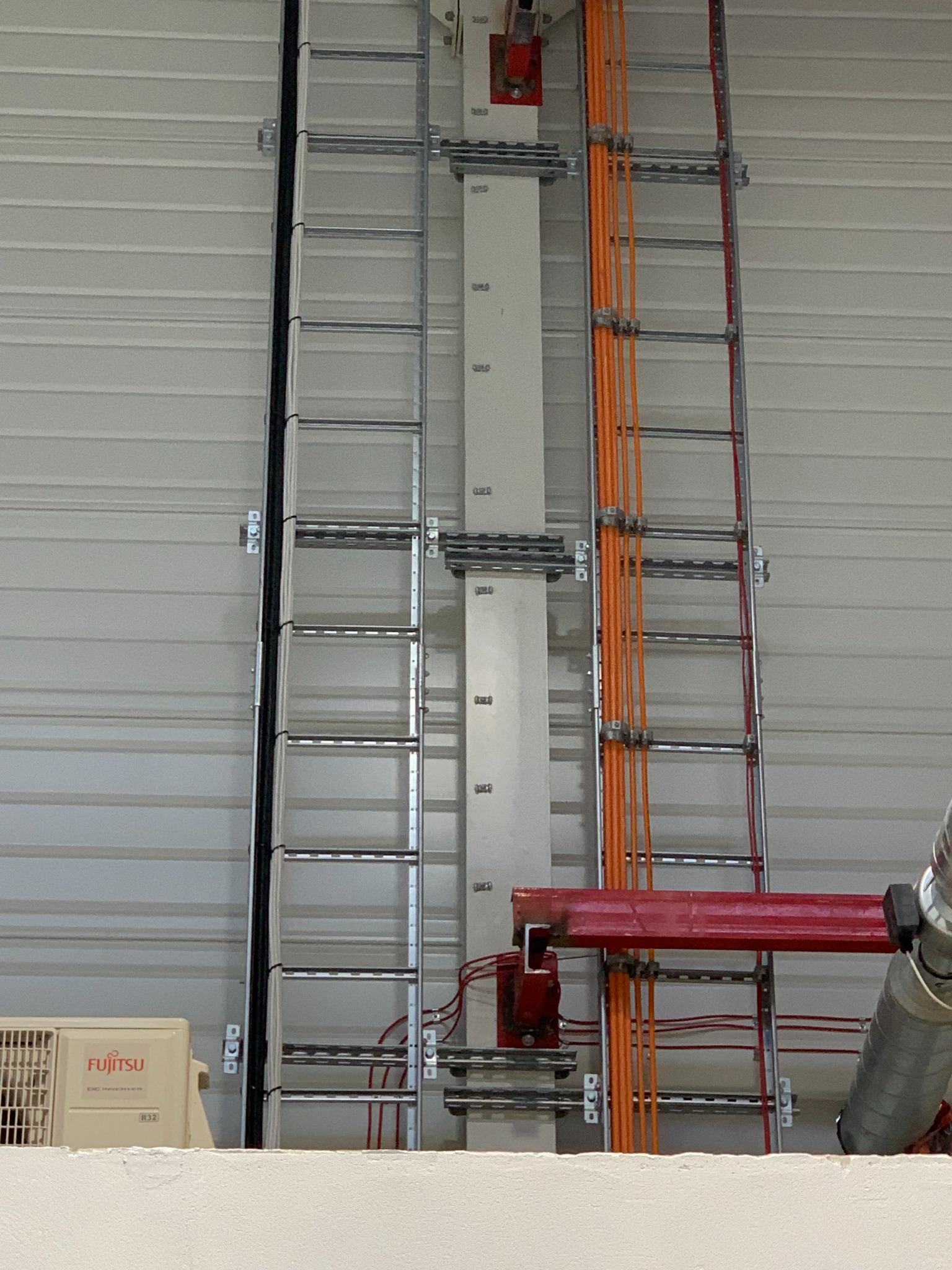 Metalowe korytka kablowe pionowo zamocowane na ścianie hali, z przewodami elektrycznymi w kolorze pomarańczowym i czarnym, widoczny klimatyzator Fujitsu.