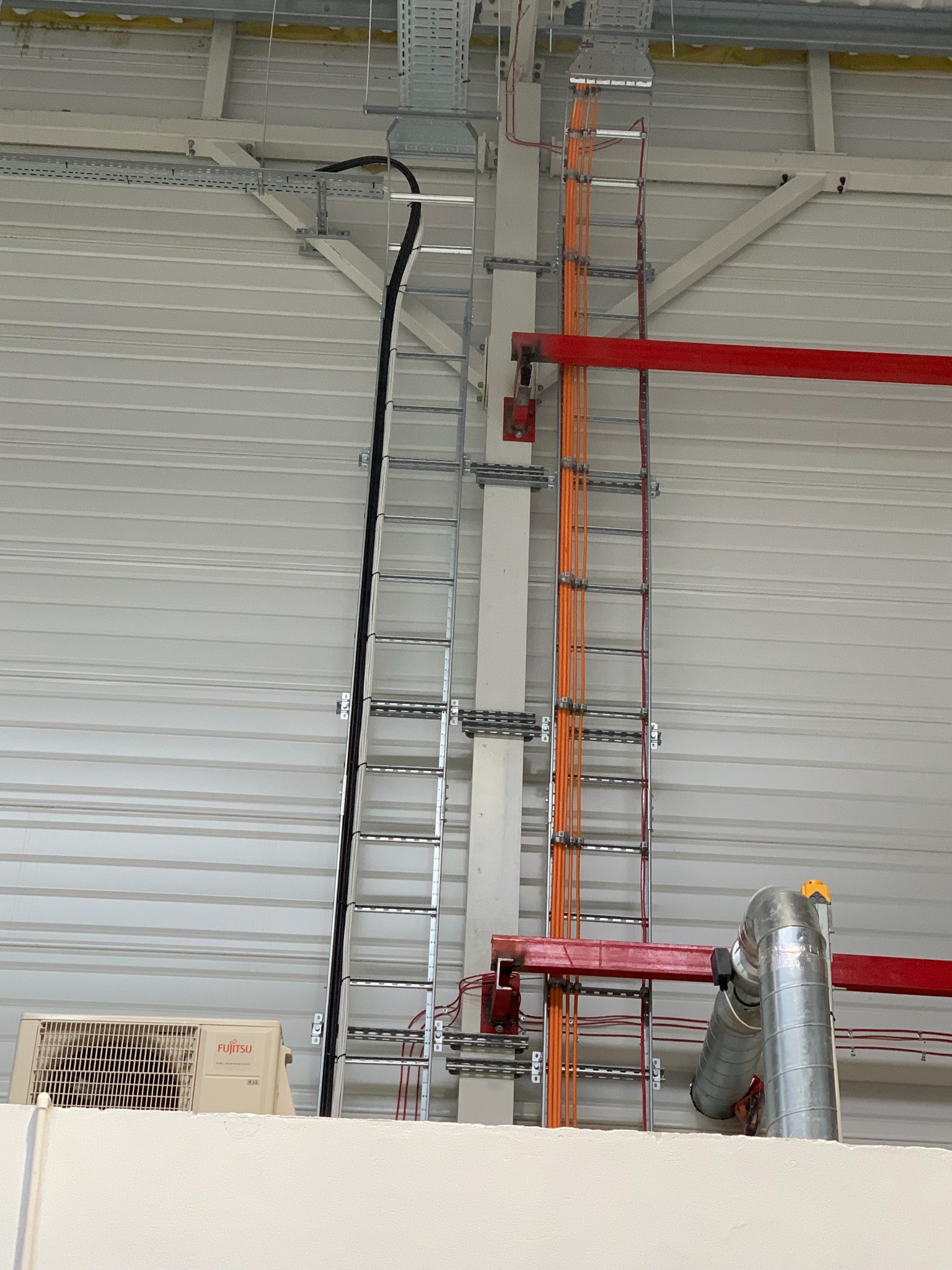 Wewnętrzna instalacja elektryczna w hali produkcyjnej: kable w korytach, rury wentylacyjne i klimatyzator Fujitsu na tle blachy trapezowej.