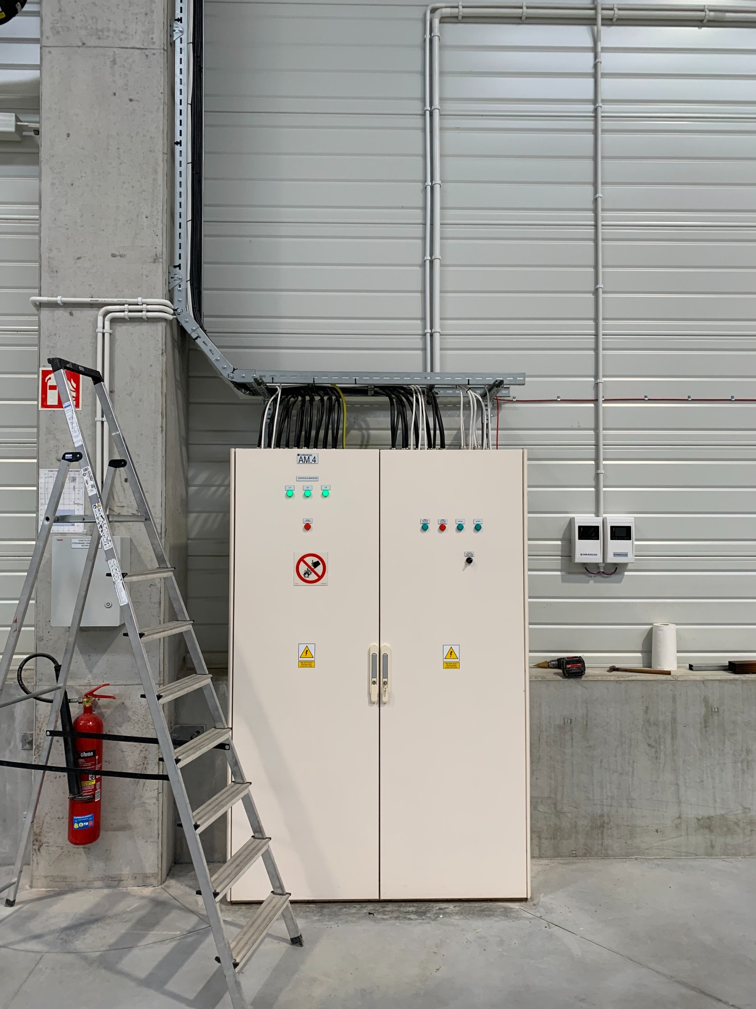 Biała szafa sterownicza z zielonymi lampkami kontrolnymi i symbolami ostrzegawczymi, zamontowana w hali przemysłowej z widoczną drabinką, gaśnicą i instalacją elektryczną pod sufitem.