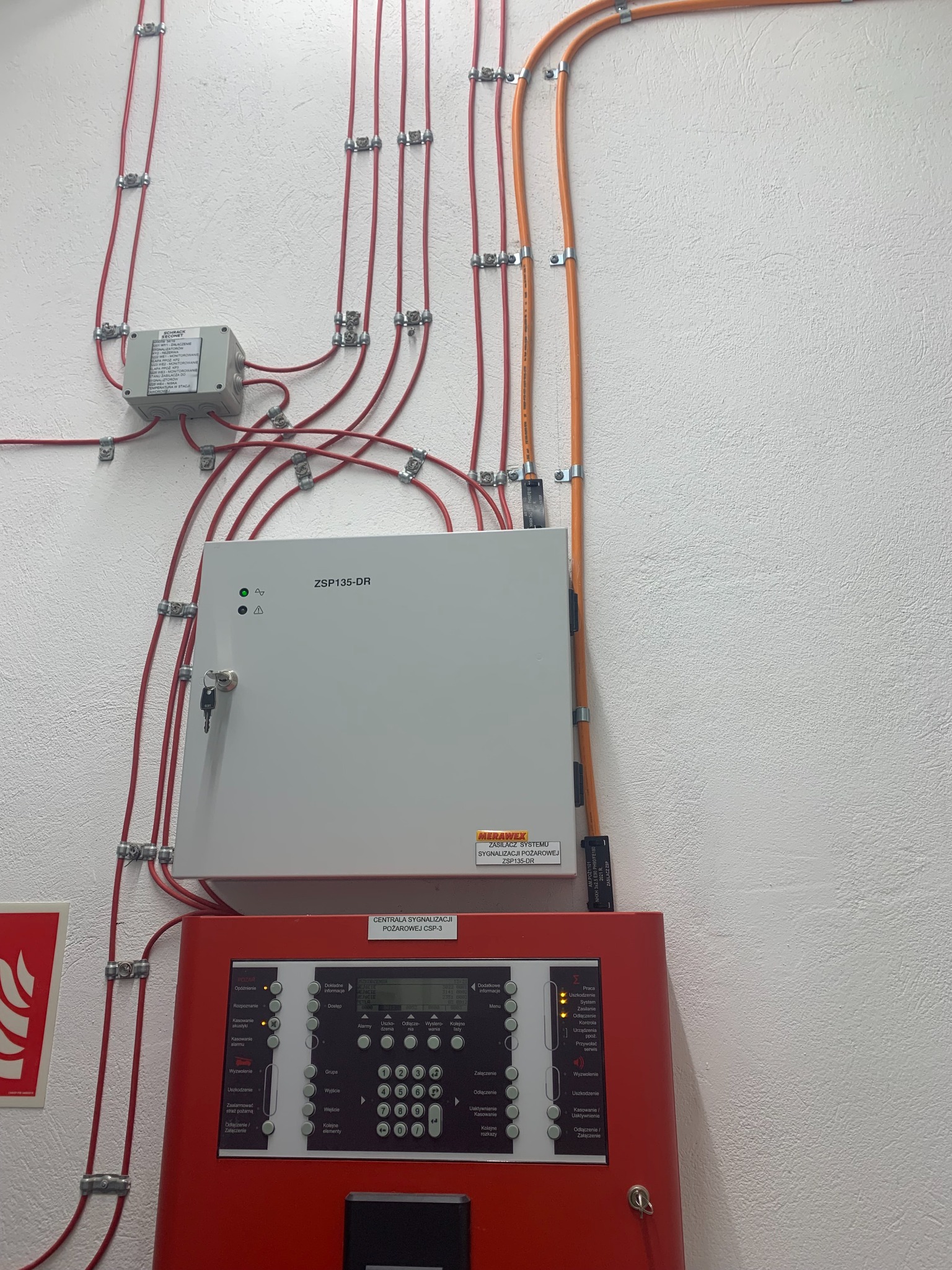 Centrala sygnalizacji pożarowej CSP-3 i zasilacz systemu sygnalizacji pożarowej ZSP135-DR na ścianie z widocznymi kablami i przewodami w metalowych uchwytach.