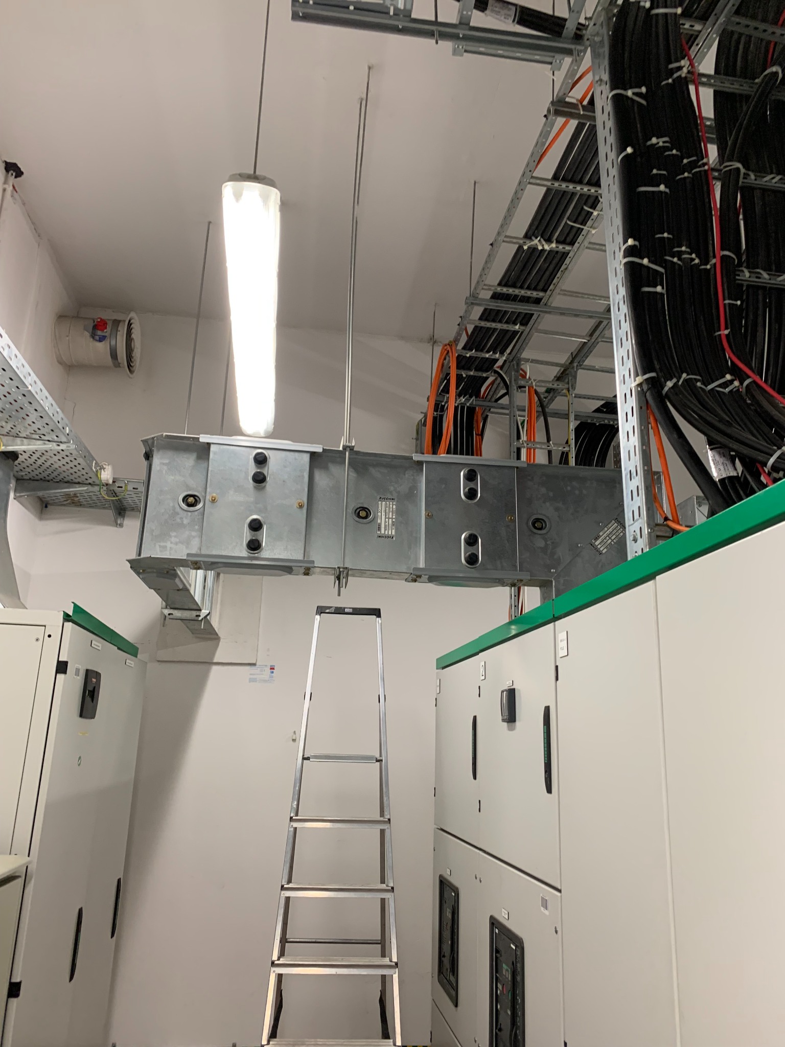Wnętrze rozdzielni elektrycznej z korytem kablowym pod sufitem, widoczna drabina aluminiowa i szafy sterownicze z zielonymi akcentami, oświetlenie jarzeniowe.