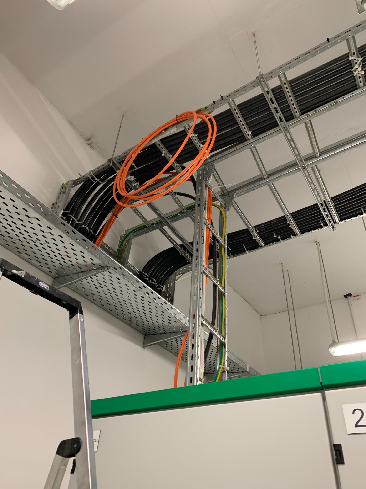 Metalowe koryta kablowe pod sufitem z ułożonymi przewodami elektrycznymi w kolorze czarnym i pomarańczowym, widoczne fragmenty drabiny i szafy sterowniczej.