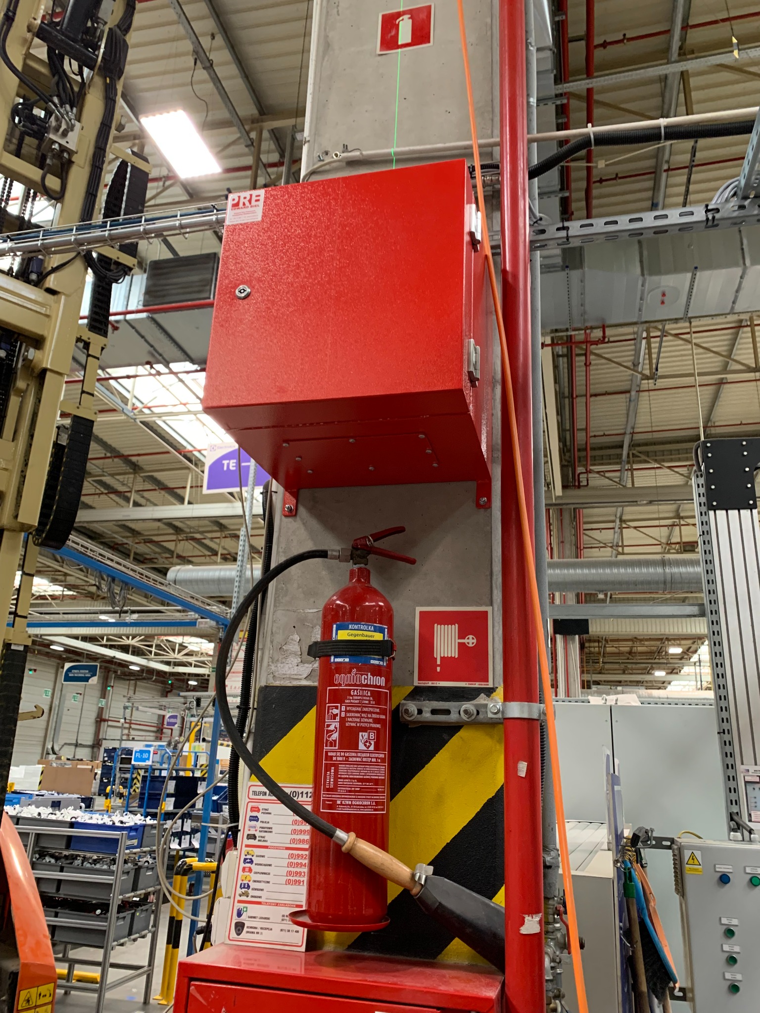 Czerwona szafka z gaśnicą w hali produkcyjnej, widoczne oznaczenia bezpieczeństwa i schemat użycia gaśnicy.
