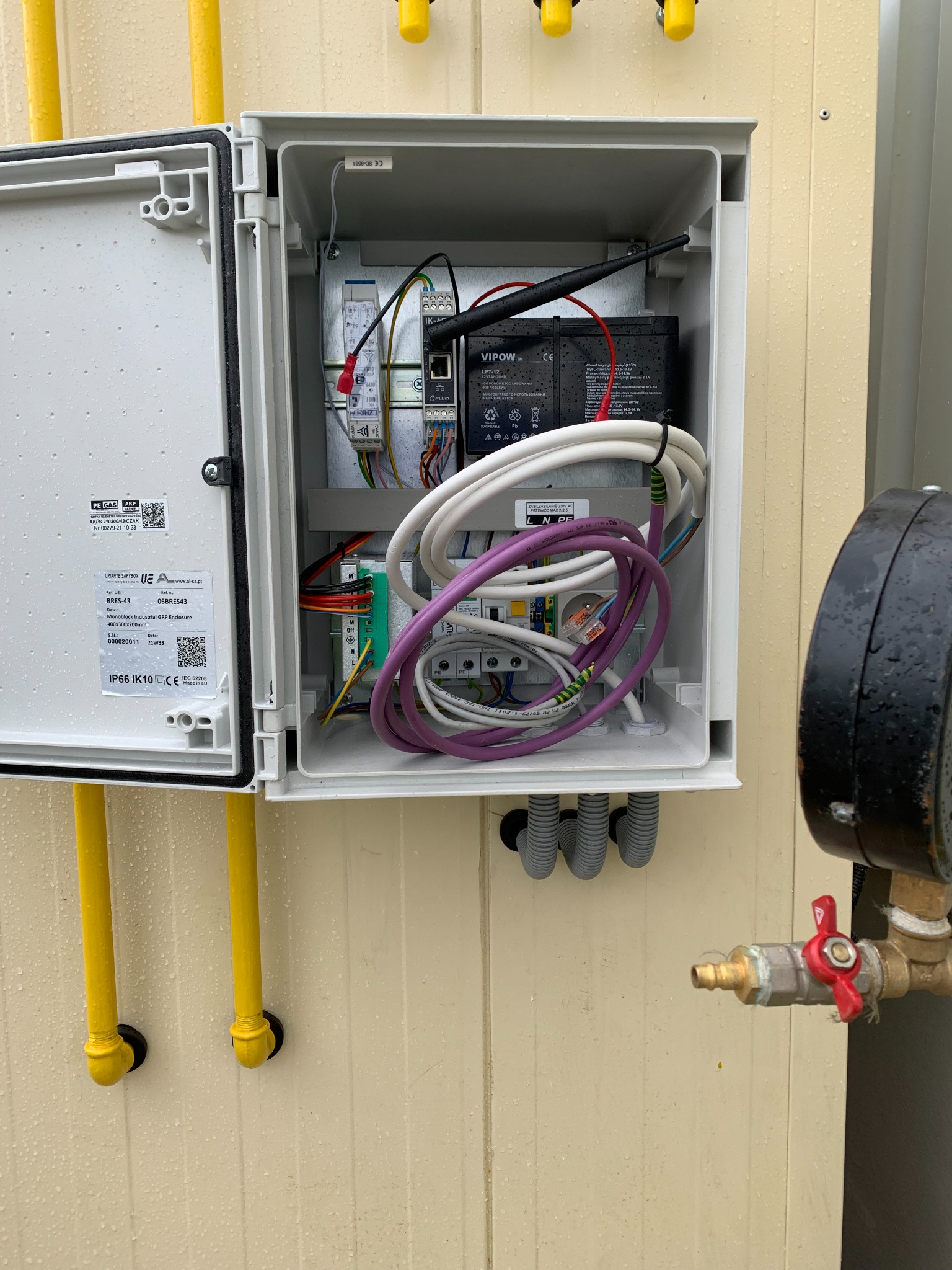 Otwarta skrzynka elektryczna z widocznymi przewodami, akumulatorem VIPOW i elementami sterującymi, zamontowana na zewnątrz budynku, obok rury gazowej z zaworem.