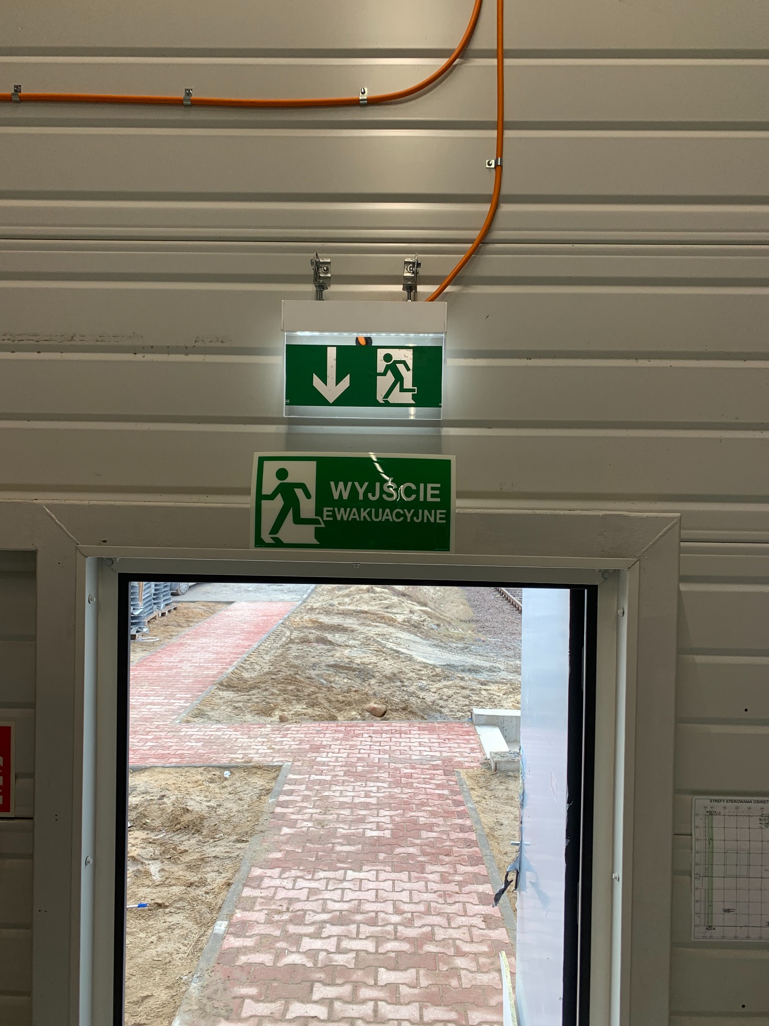 Znak 'Wyjście Ewakuacyjne' z podświetleniem LED, zamontowany nad drzwiami wewnątrz budynku o ścianach z blachy trapezowej, widoczny pomarańczowy przewód elektryczny w metalowych uchwytach.
