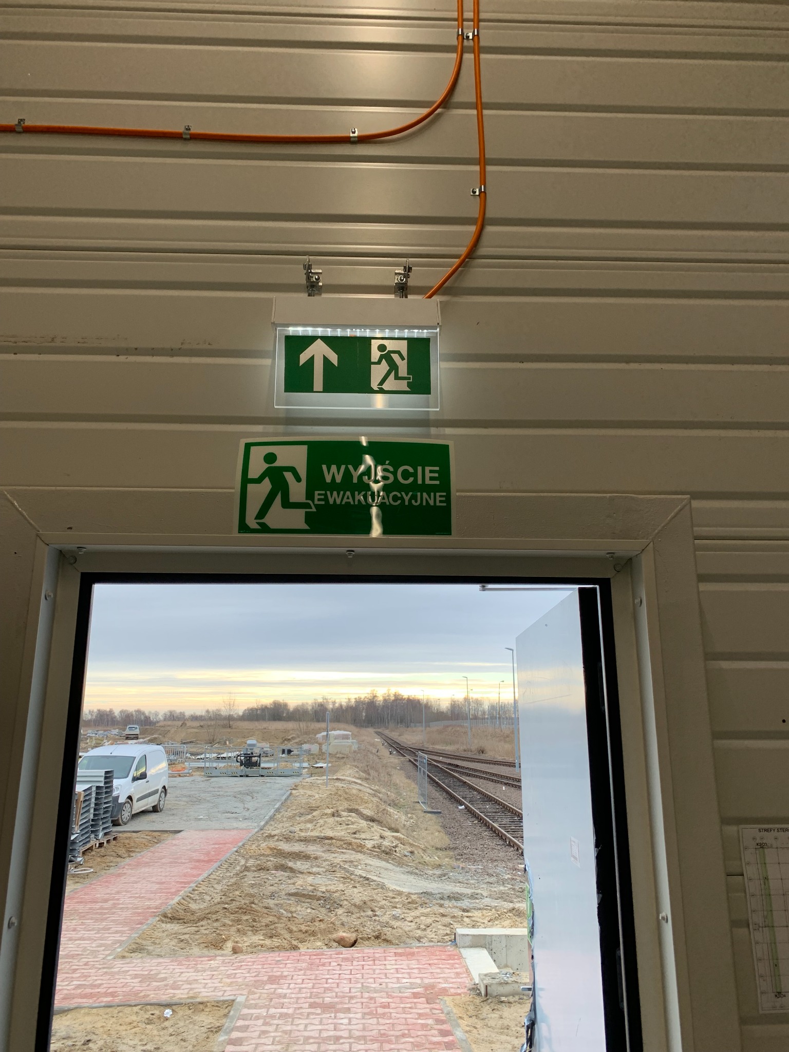 Wewnętrzne oznaczenia drogi ewakuacyjnej z podświetlanym znakiem, widok z pomieszczenia na plac budowy z ułożoną kostką, widoczna instalacja elektryczna w pomarańczowej rurce.