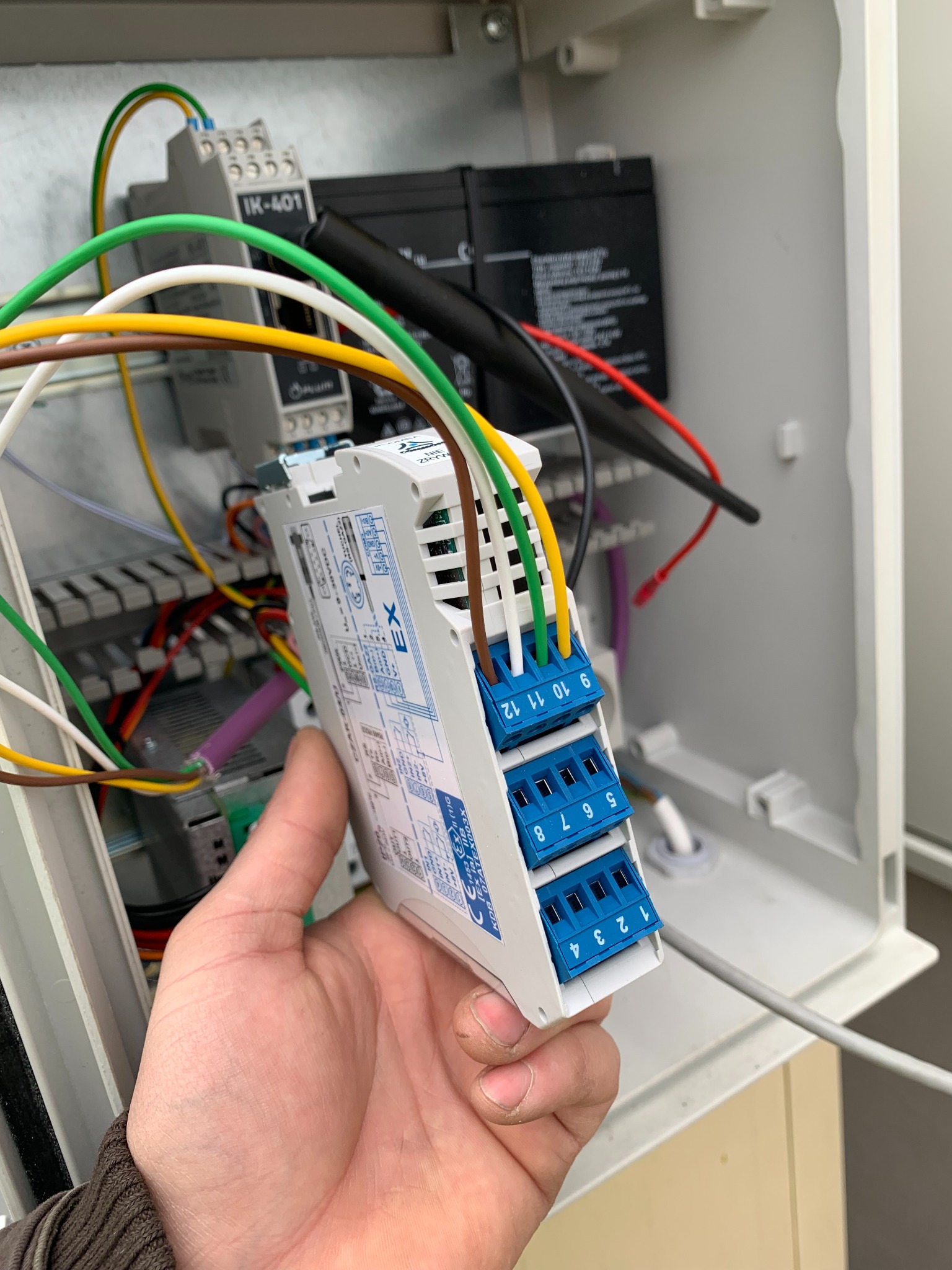 Ręka trzyma moduł sterujący z niebieskimi złączami i podłączonymi kolorowymi przewodami wewnątrz skrzynki elektrycznej, widoczne akumulatory i inne elementy instalacji.
