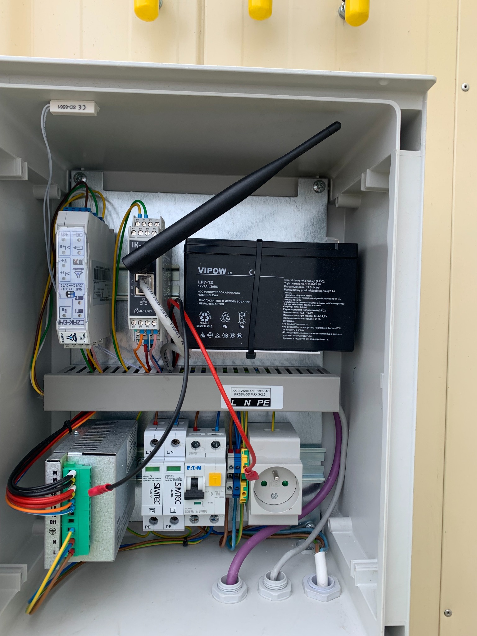 Wnętrze skrzynki elektrycznej z akumulatorem VIPOW, modułami SIMTEC, zasilaczem, gniazdkiem i anteną WiFi, okablowanie uporządkowane, widoczne elementy instalacji alarmowej lub zasilania awaryjnego.