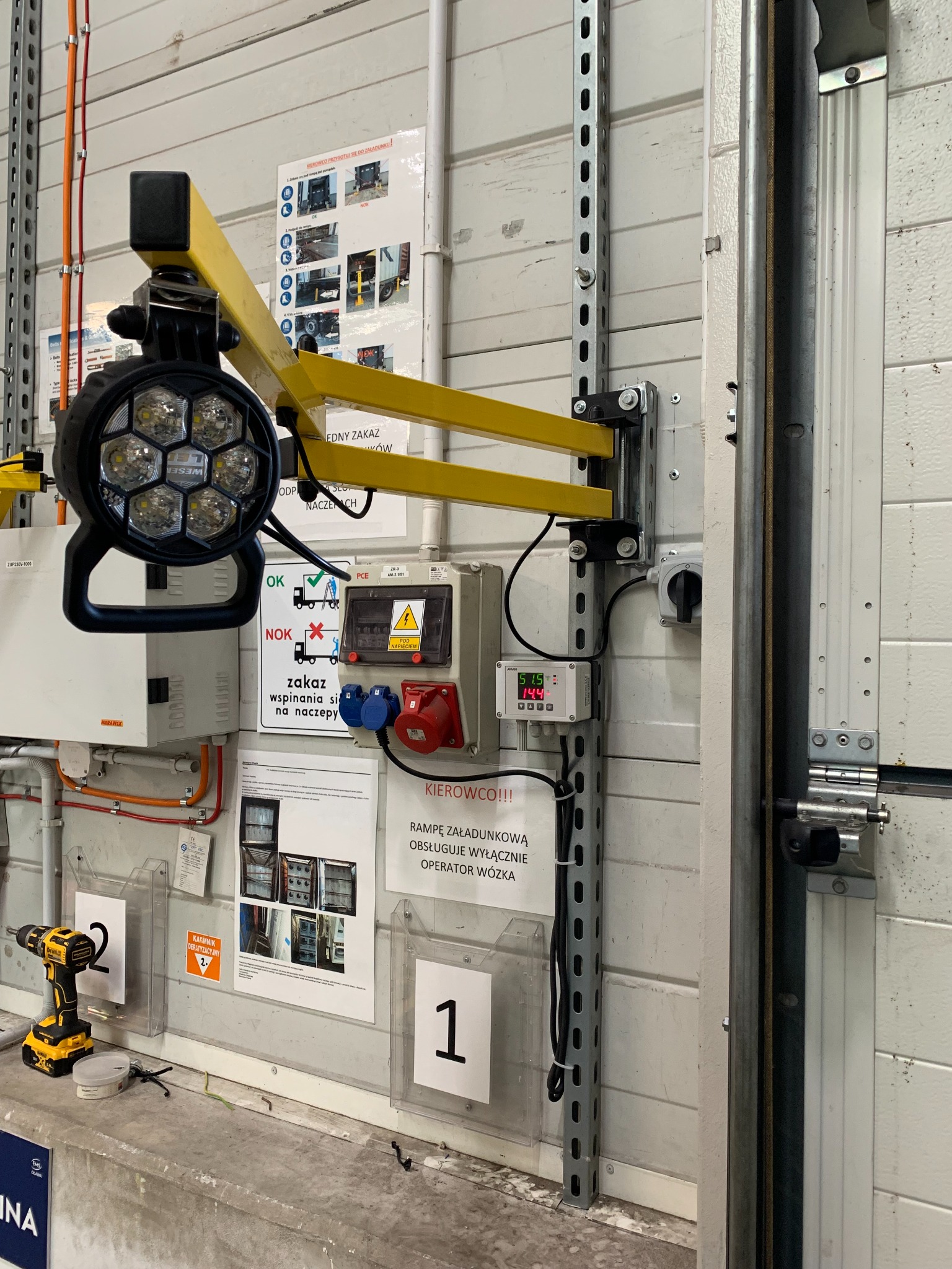 Wewnętrzne oświetlenie LED na wysięgniku zamontowane na szynie zasilającej, obok skrzynki elektrycznej, gniazd i wyświetlacza, z wiertarką akumulatorową leżącą obok na betonowej posadzce w hali...