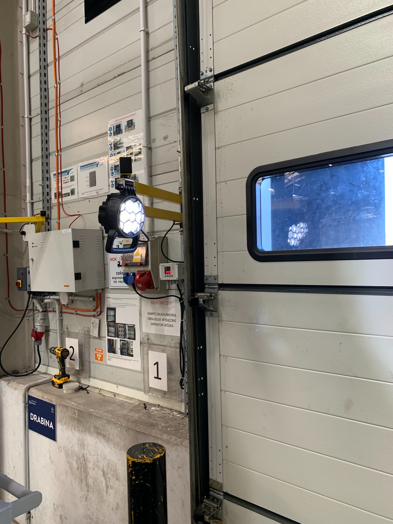 Oświetlenie rampy załadunkowej z panelem sterowania, gniazdami zasilającymi i instrukcjami obsługi na ścianie hali, obok otwartej bramy segmentowej z oknem. Widoczna wkrętarka akumulatorowa i znak...