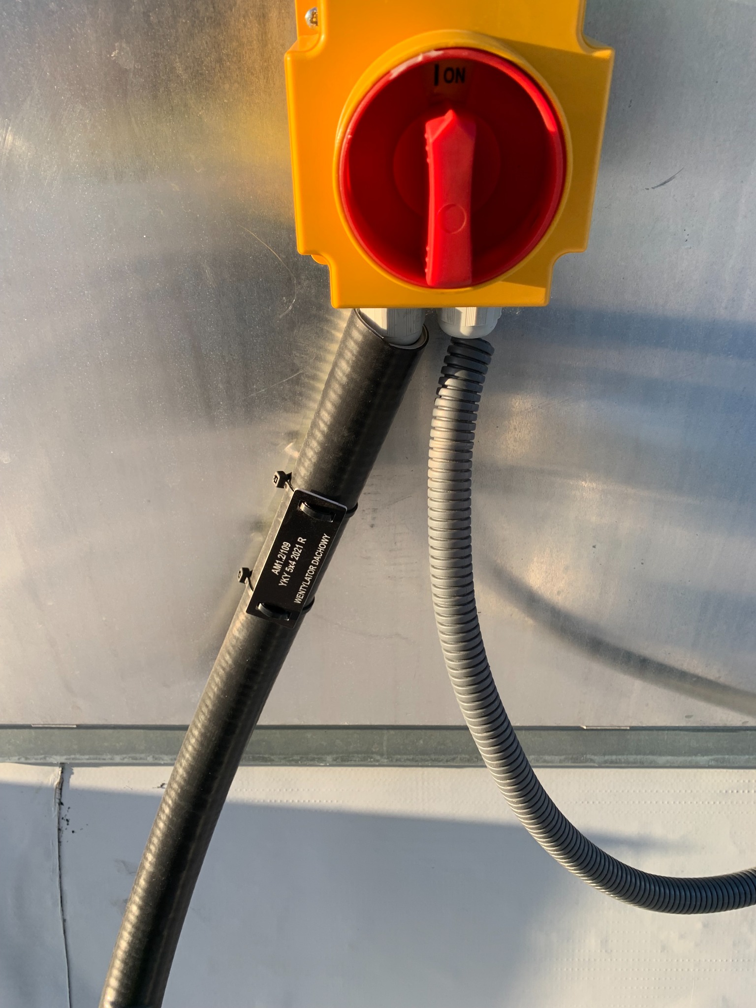 Żółty wyłącznik bezpieczeństwa z czerwoną dźwignią w pozycji ON, przymocowany do metalowej powierzchni, z podłączonymi przewodami w peszlach, jeden z etykietą 'WENTYLATOR DACHOWY'.