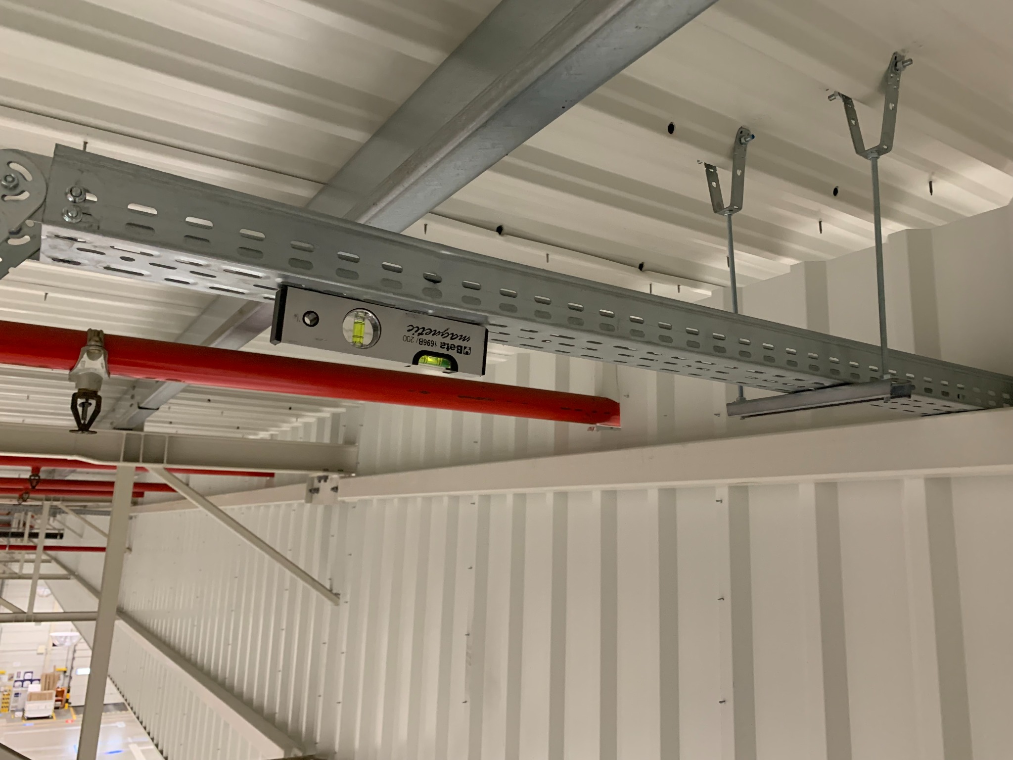 Montaż perforowanej, metalowej korytki kablowej pod sufitem hali przemysłowej, poziomowana za pomocą poziomicy magnetycznej, widoczna instalacja przeciwpożarowa.