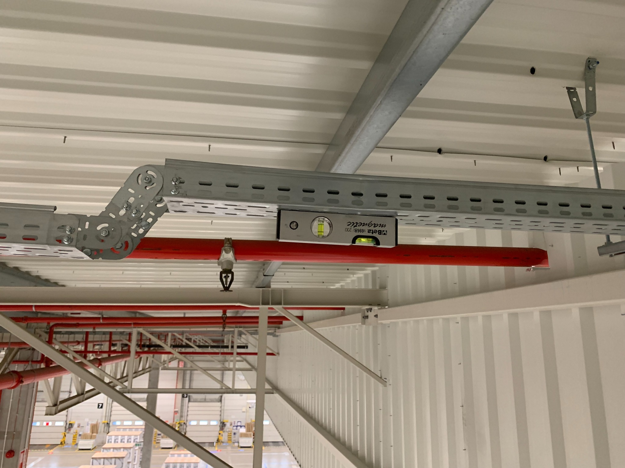 Poziomica magnetyczna na metalowej konstrukcji wsporczej w hali magazynowej w Wysokiej, widoczne rury instalacji przeciwpożarowej i fragmenty konstrukcji dachu.