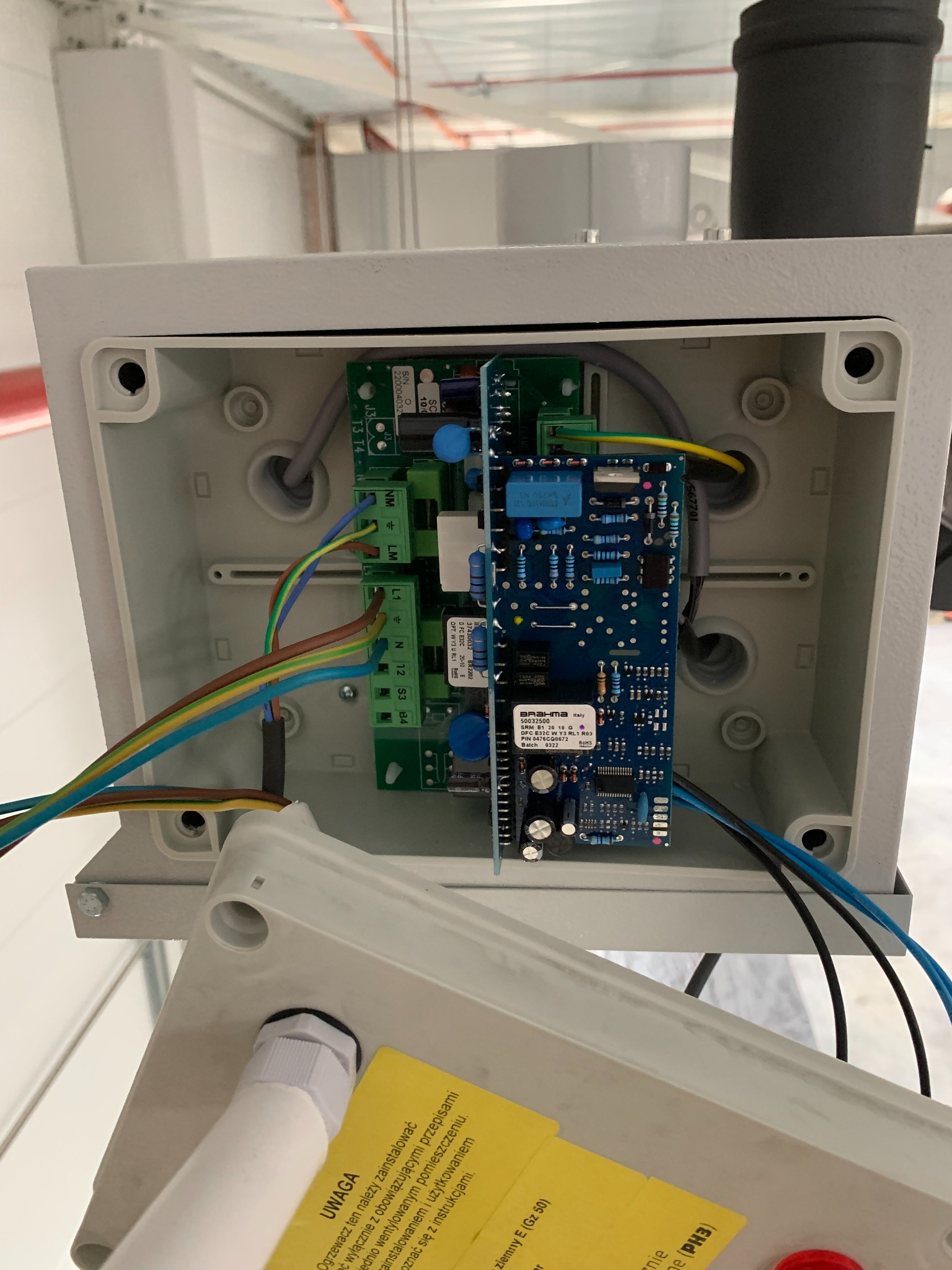 Otwarta, szara skrzynka elektryczna z widocznymi płytkami drukowanymi, przewodami w różnych kolorach i żółtą naklejką ostrzegawczą z tekstem w języku polskim, zamontowana w przemysłowym otoczeniu.