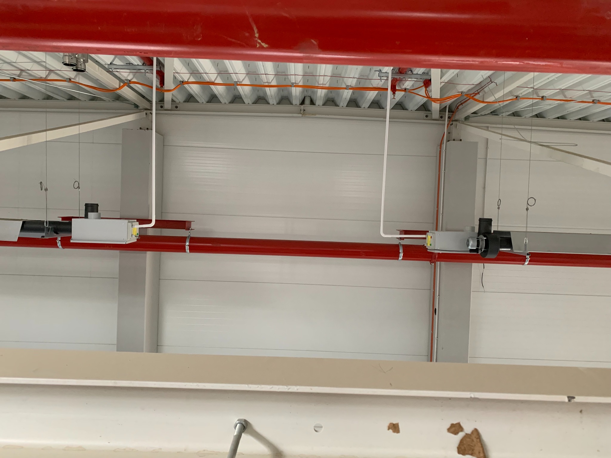 Widok instalacji pod sufitem w hali przemysłowej z widocznymi kablami elektrycznymi, rurami w kolorze czerwonym oraz elementami systemu wentylacji.