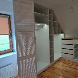 Biała szafa wnękowa z drewnianymi lamelowymi drzwiami przesuwnymi, wkomponowana w skos poddasza, oświetlenie LED wewnątrz, parkiet na podłodze.