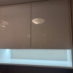 WOOD ART. MEBLE RAFAŁ CHALECKI - Biała, lakierowana szafka wisząca z oświetleniem LED podszafkowym, umieszczona nad blatem w kuchni, widoczna lampa sufitowa.