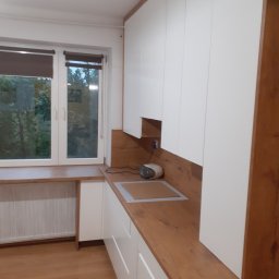 WOOD ART. MEBLE RAFAŁ CHALECKI - Biała zabudowa kuchenna z drewnianym blatem i okładziną ścienną, widok na okno z blatem, zlew wpuszczony w blat, małe radio na blacie.