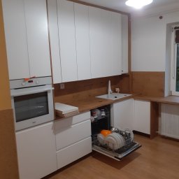 WOOD ART. MEBLE RAFAŁ CHALECKI - Biała kuchnia na wymiar z drewnianym blatem i otwartą zmywarką wypełnioną naczyniami, widoczne także piekarnik i okno.