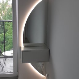 WOOD ART. MEBLE RAFAŁ CHALECKI - Biała toaletka wisząca z szufladą i podświetleniem LED w kształcie półksiężyca, widok z boku na tle okna balkonowego i fragmentu łóżka.