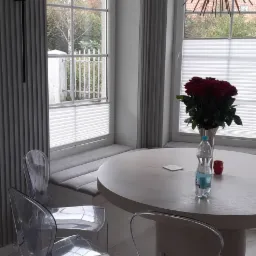 Jasne wnętrze jadalni z okrągłym stołem, bukietem czerwonych róż i przezroczystymi krzesłami; w tle okno z roletami plisowanymi i siedziskiem wnękowym.