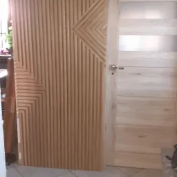 Drewniana ściana działowa z geometrycznym wzorem w pionowe lamele, sąsiadująca z jasnymi, drewnianymi drzwiami z przeszkleniem w górnej części; widoczne fragmenty otoczenia.