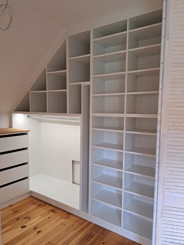 Biała zabudowa wnęki na poddaszu ze schowkiem i regałem z półkami, widoczna drewniana podłoga.