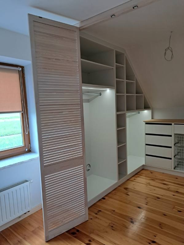 Biała szafa wnękowa z drewnianymi lamelowymi drzwiami przesuwnymi, wkomponowana w skos poddasza, oświetlenie LED wewnątrz, parkiet na podłodze.