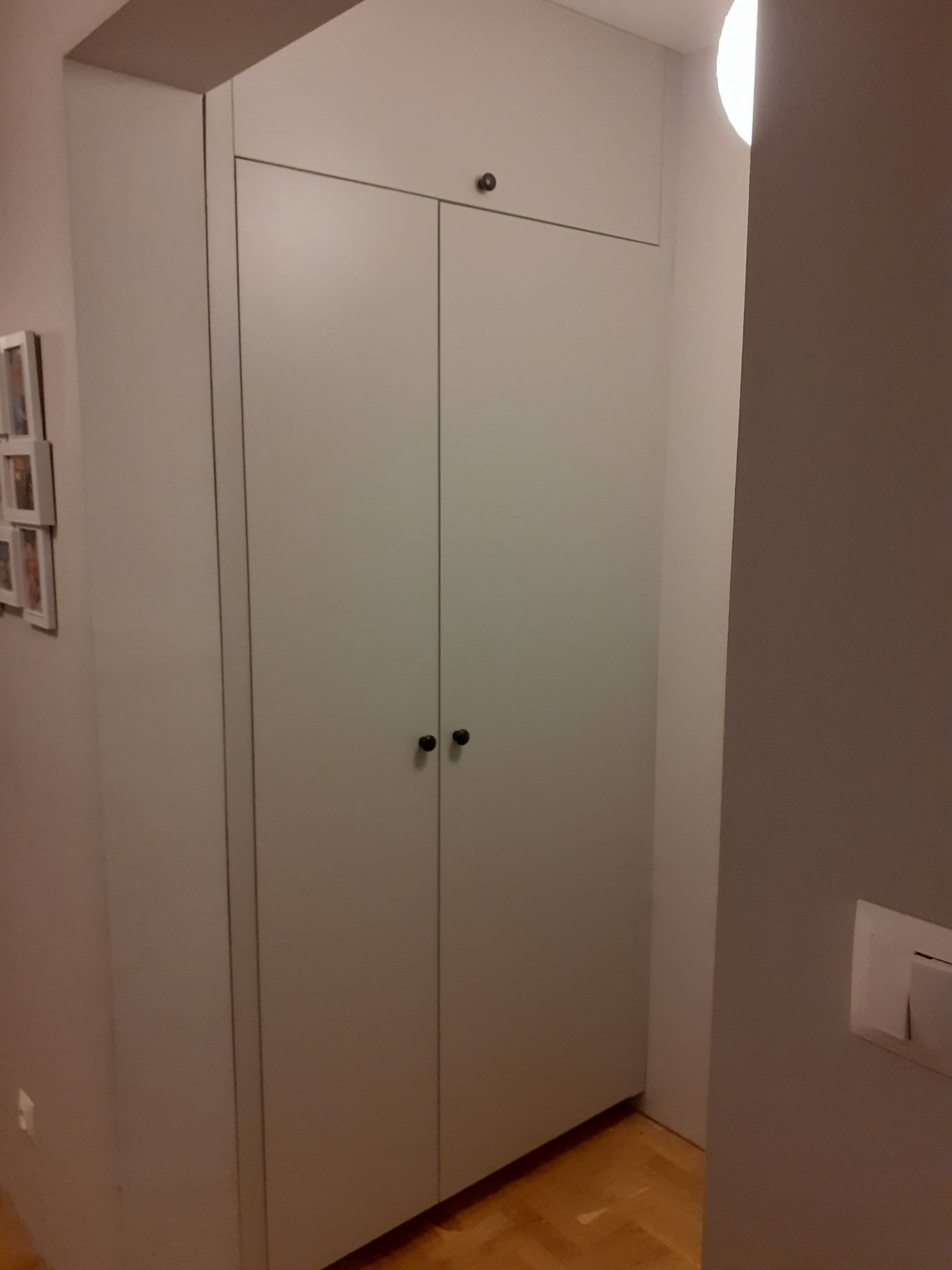 Wysoka, wbudowana szafa z jasnymi, gładkimi frontami i małymi, okrągłymi uchwytami, umieszczona w przedpokoju z parkietem i dekoracjami na ścianie.