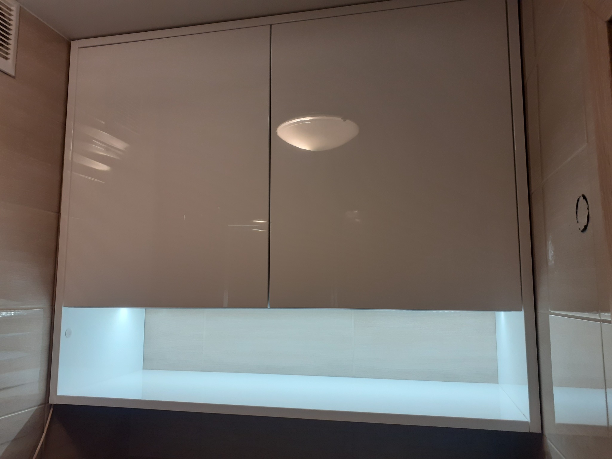 Biała, lakierowana szafka wisząca z oświetleniem LED podszafkowym, umieszczona nad blatem w kuchni, widoczna lampa sufitowa.