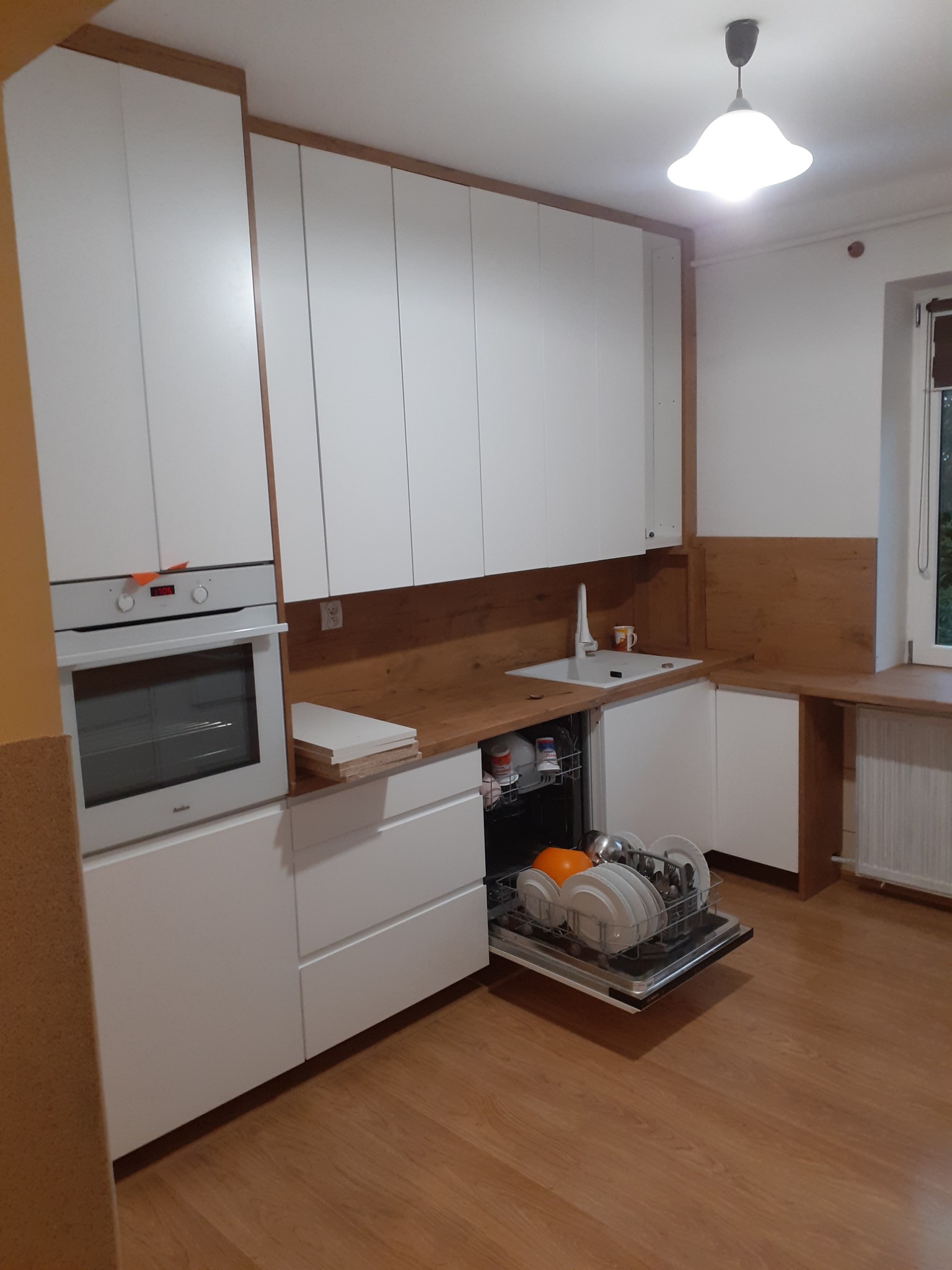 Biała kuchnia na wymiar z drewnianym blatem i otwartą zmywarką wypełnioną naczyniami, widoczne także piekarnik i okno.