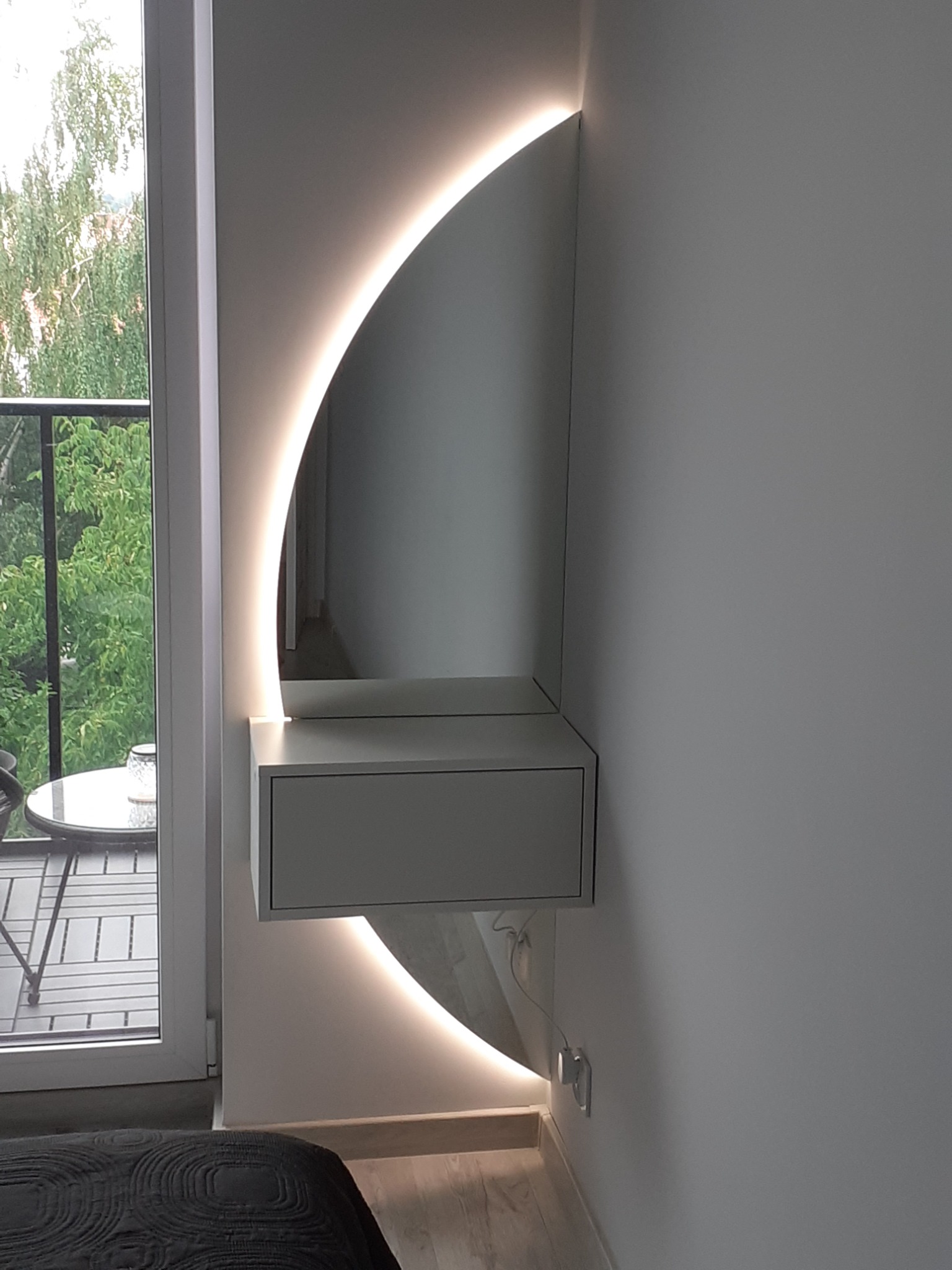 Biała toaletka wisząca z szufladą i podświetleniem LED w kształcie półksiężyca, widok z boku na tle okna balkonowego i fragmentu łóżka.